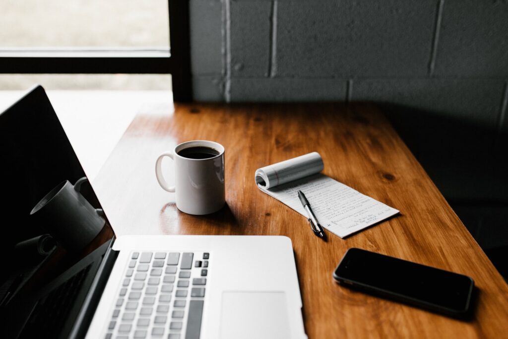 
A notepad, pen, coffee mug, laptop, phone on a wooden table