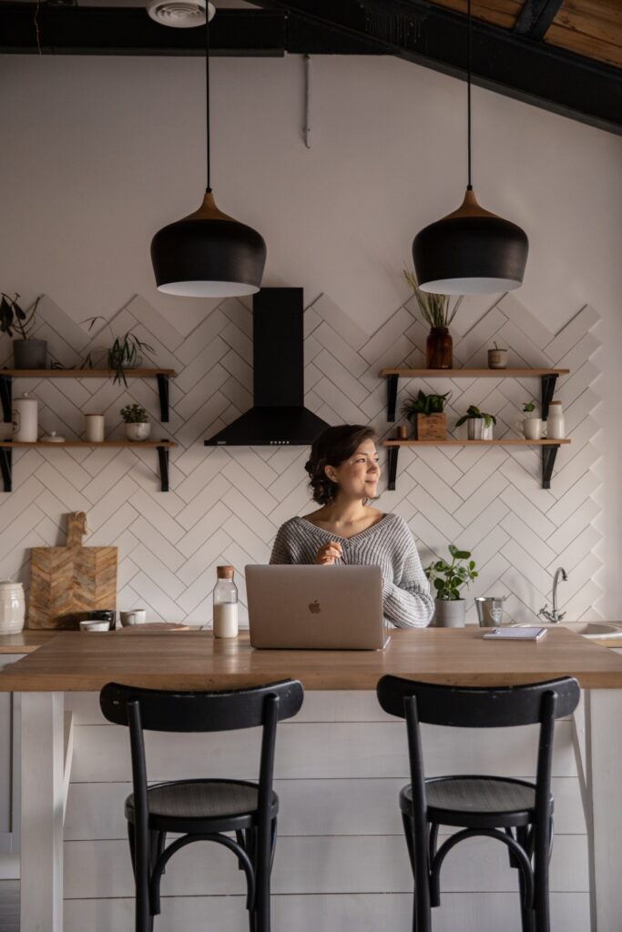 A lady looking out of the window. She is sitting at the kitchen table , working on her laptop.