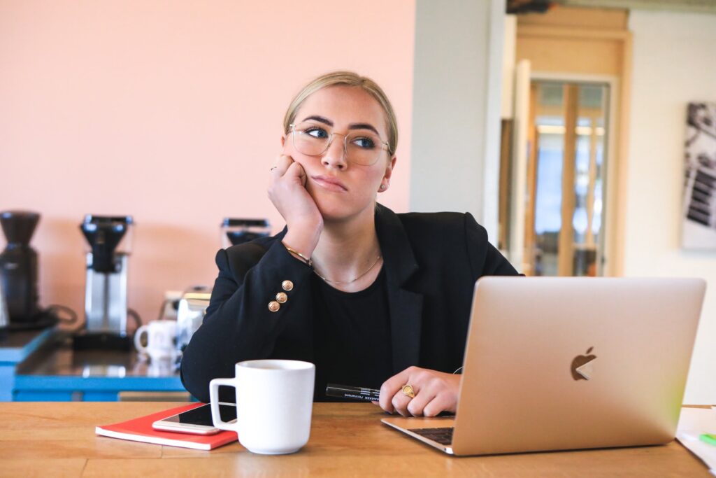 A lady thinking in front of her laptop with a book and coffee mug at the table